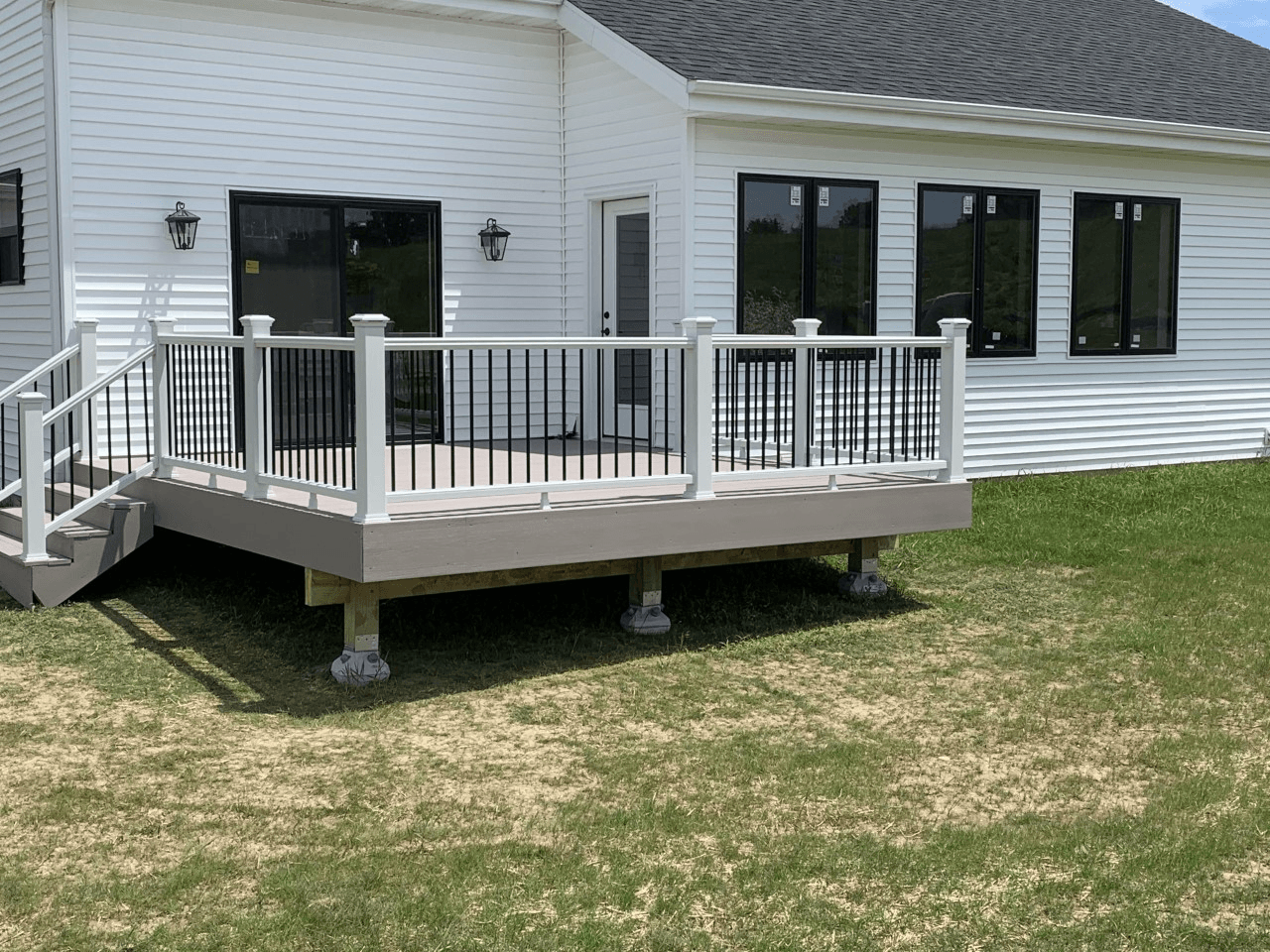 New deck attached to white house with black framed windows. Gray deck, black railing, gray support columns on grass.