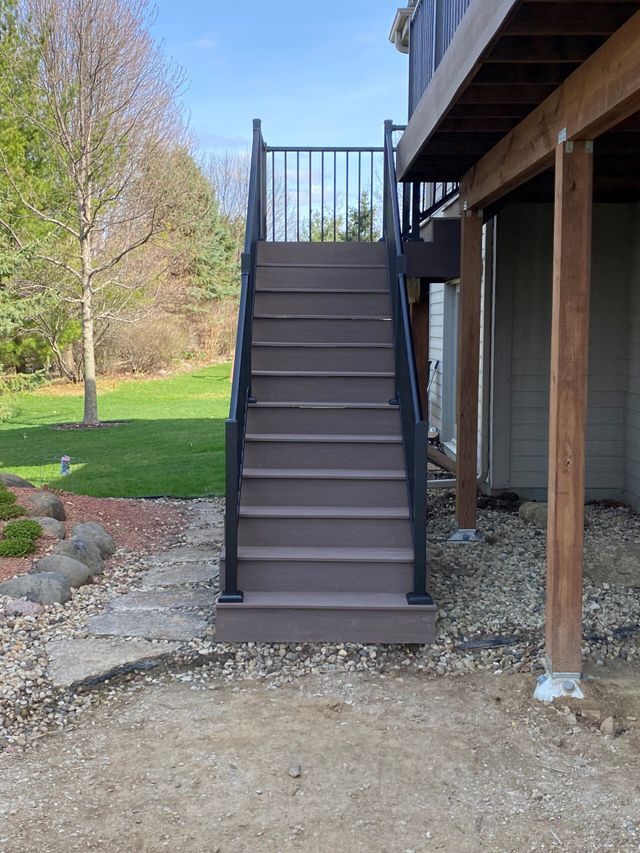 Staircase leading up to a deck. Brown composite steps with black metal railing, gravel and grass surroundings.
