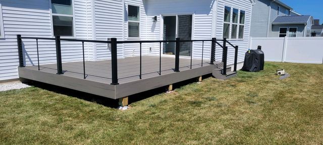 A backyard deck with black railing and steps is next to a house with white siding and a grassy lawn.