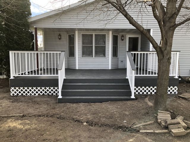 Gray deck with white railing, steps leading to a light gray house with windows. Bare tree in foreground.