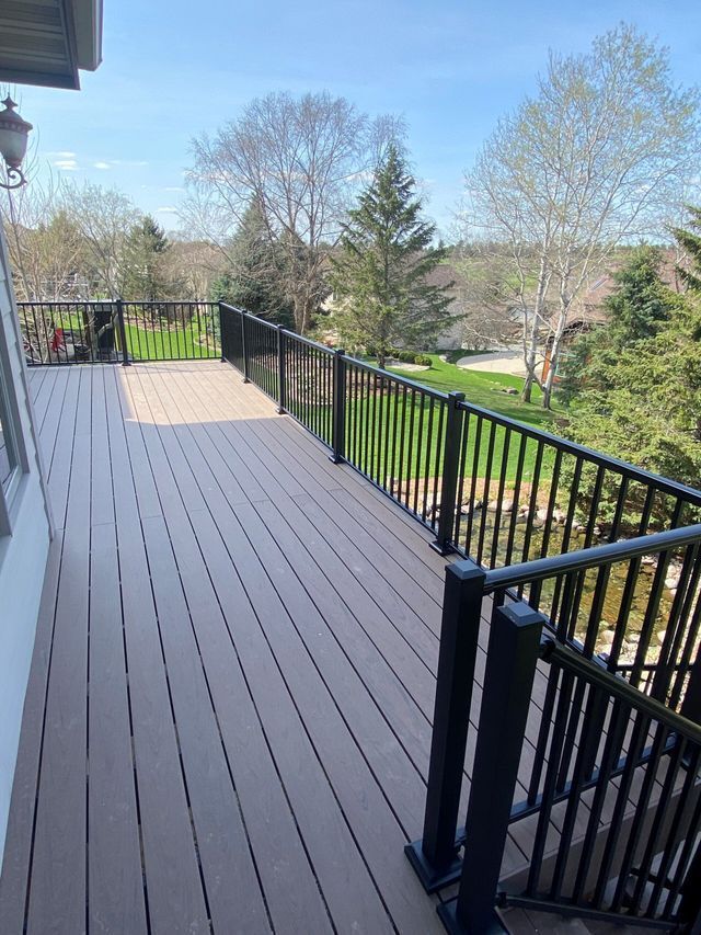 Wooden deck with black railings overlooking a green lawn and trees under a blue sky.