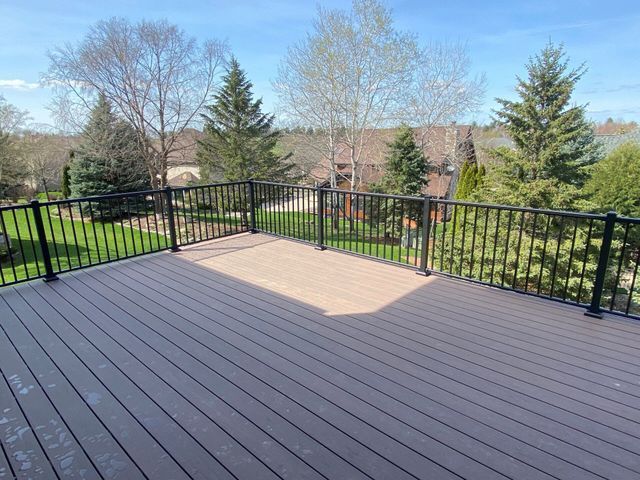 A composite deck with black railing overlooks a grassy backyard and trees under a blue sky.
