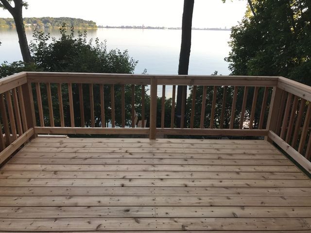 Wooden deck overlooking a calm lake, surrounded by trees and a railing.