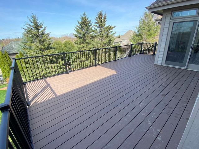 Brown composite deck with black metal railing, overlooking green trees and a blue sky.