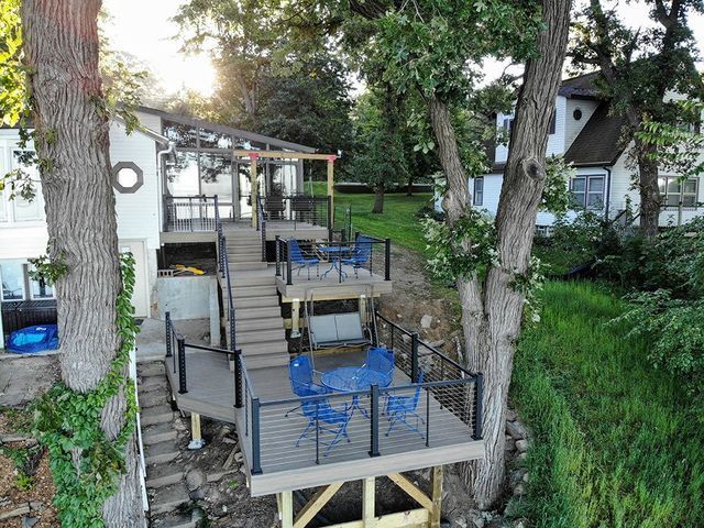 Multi-level deck on a white house, with blue chairs and black railings. Trees frame the scene near green grass.