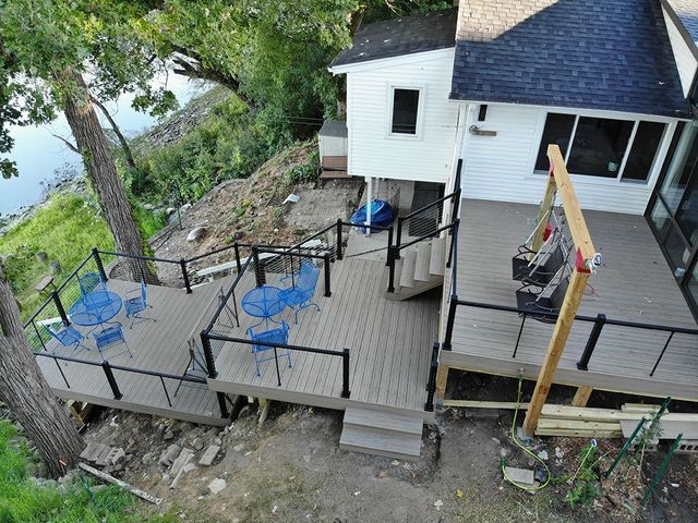 Multi-level deck with blue chairs, overlooking water. White house, trees, and black railing.