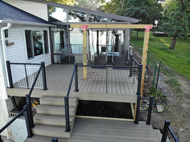 Deck with steps, railing, and a pergola-like structure extending from a house, overlooking a grassy area.