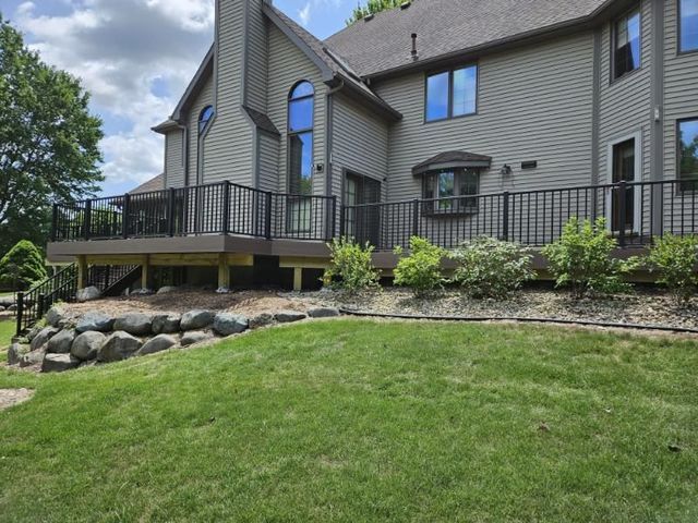 Back of a house with a large deck, black railing, and a rock retaining wall along the edge of the lawn.