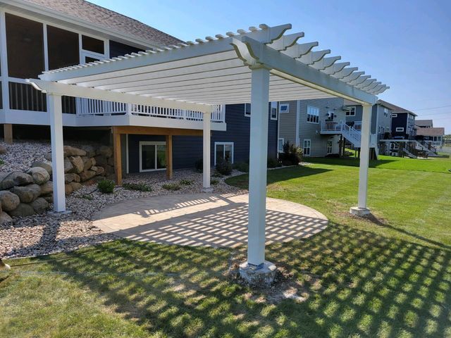White pergola over a concrete patio with shadows on grass, near a house.
