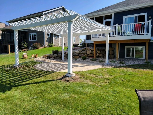 White pergola over patio in a backyard with a blue house, rock wall, and green lawn.