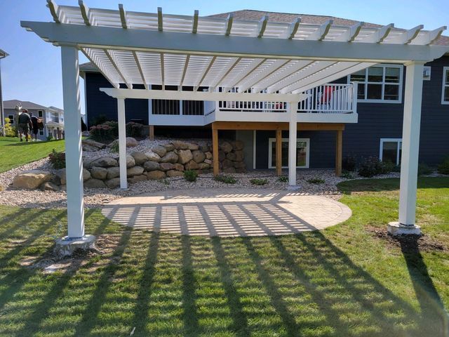 White pergola over a concrete patio casting shadows on green grass, next to a house with a deck.
