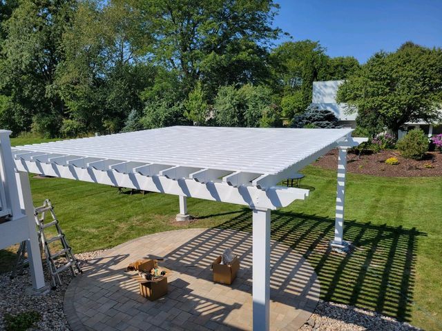 White pergola over a patio, casting shadows on the stone and lawn, with trees in the background.