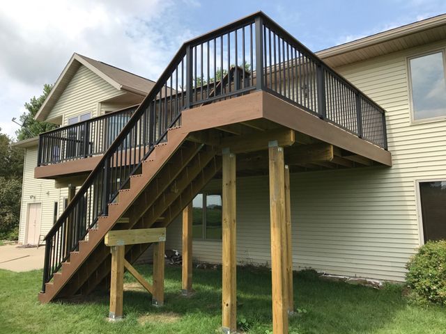 A two-story deck with black railings and brown decking, supported by wooden posts, next to a tan house.