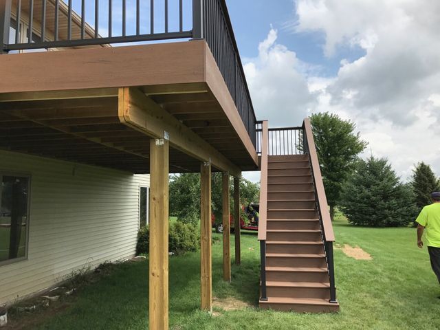 A two-level brown deck with black railing and stairs in a grassy yard under a cloudy sky.
