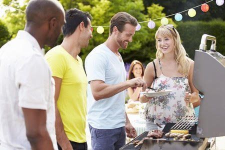 Group of people gathered around a grill outdoors; smiling, interacting, and enjoying a barbecue.