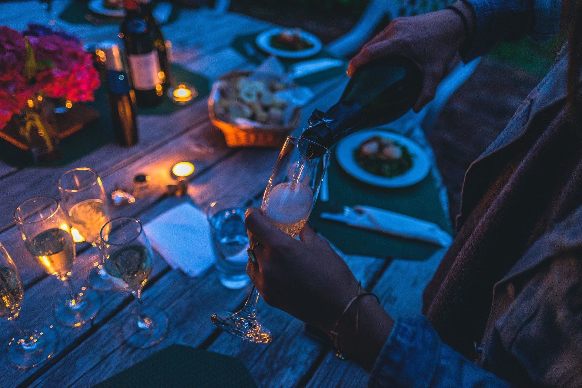 Person pouring champagne into a glass at an outdoor table with food, drinks, and candles.