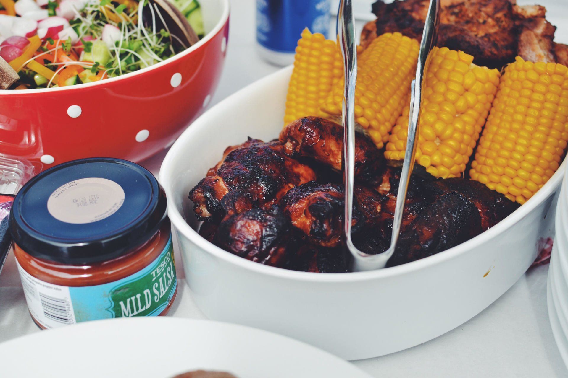 Barbecued ribs, corn on the cob, and salad in a red bowl with sauce on a white table.