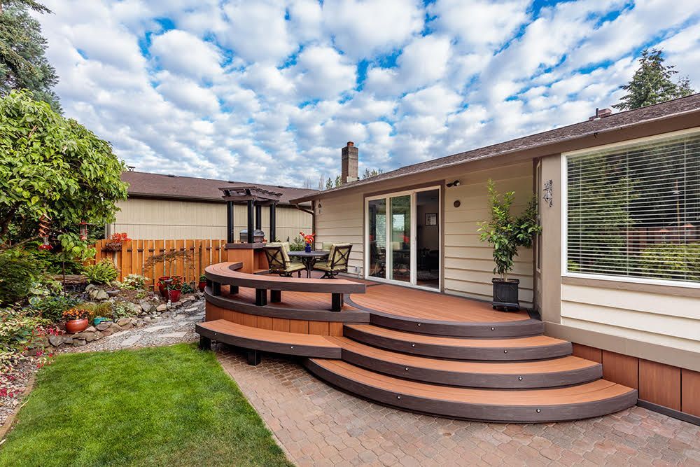 Wooden deck with curved steps, leading to a house with a large window and sliding glass door, surrounded by a yard and garden.