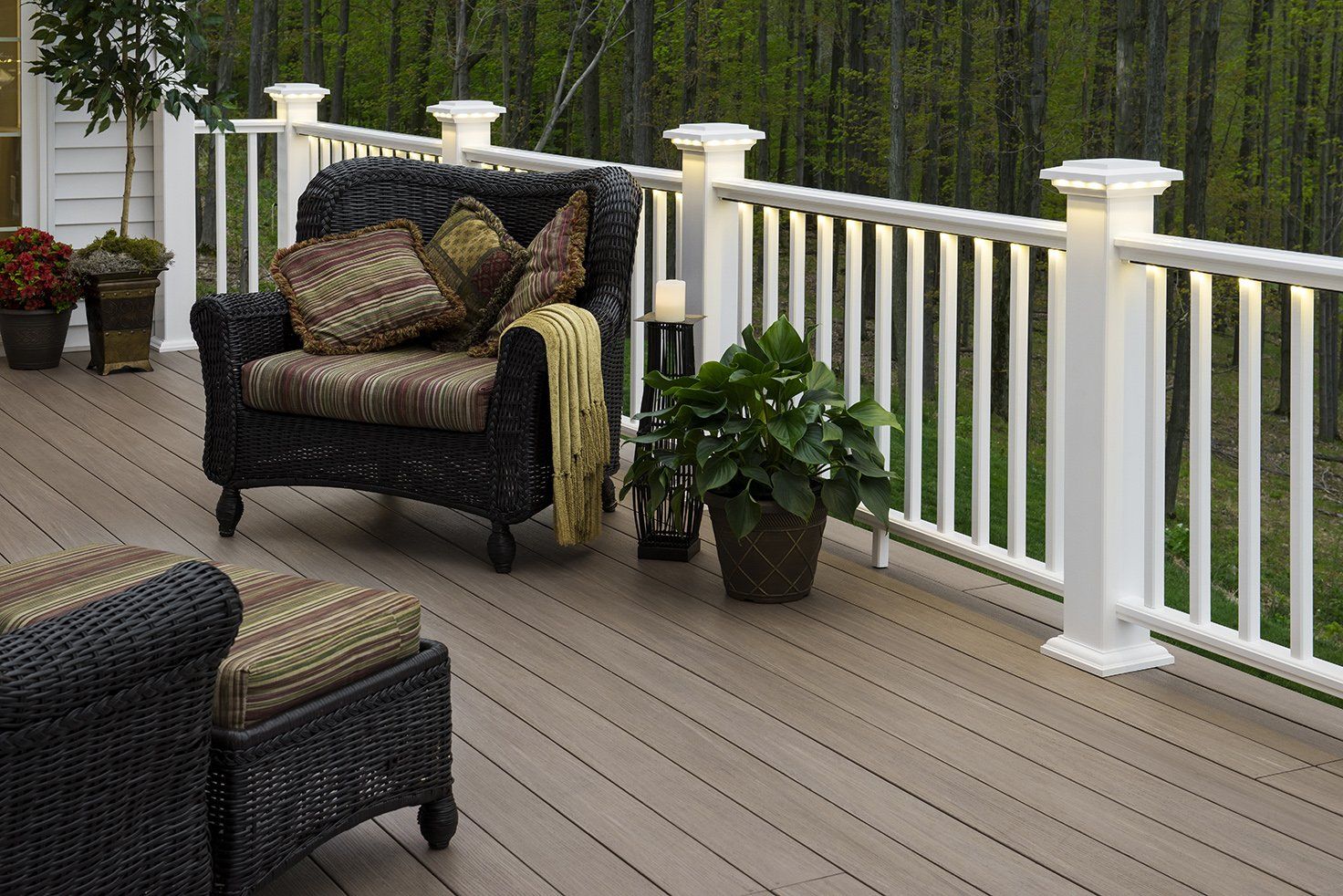 Wicker furniture on a deck with white railings and lights, overlooking a wooded area.