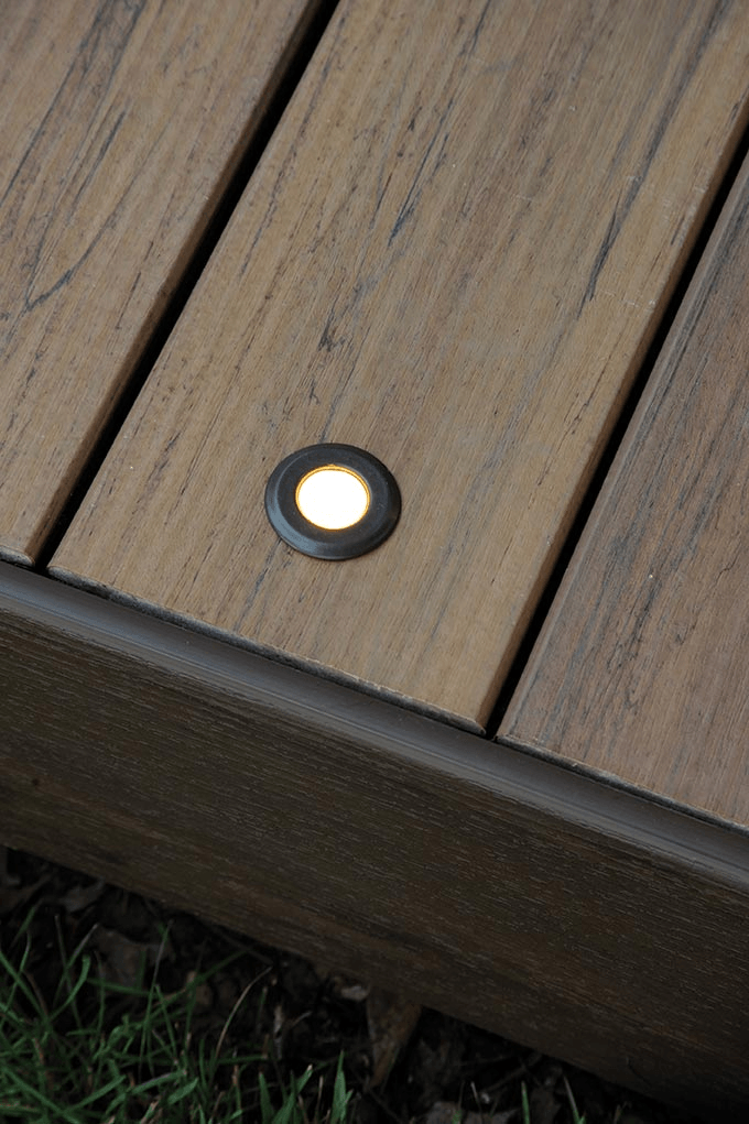 Close-up of a deck with a recessed, illuminated light. The deck is brown, and the light is glowing.