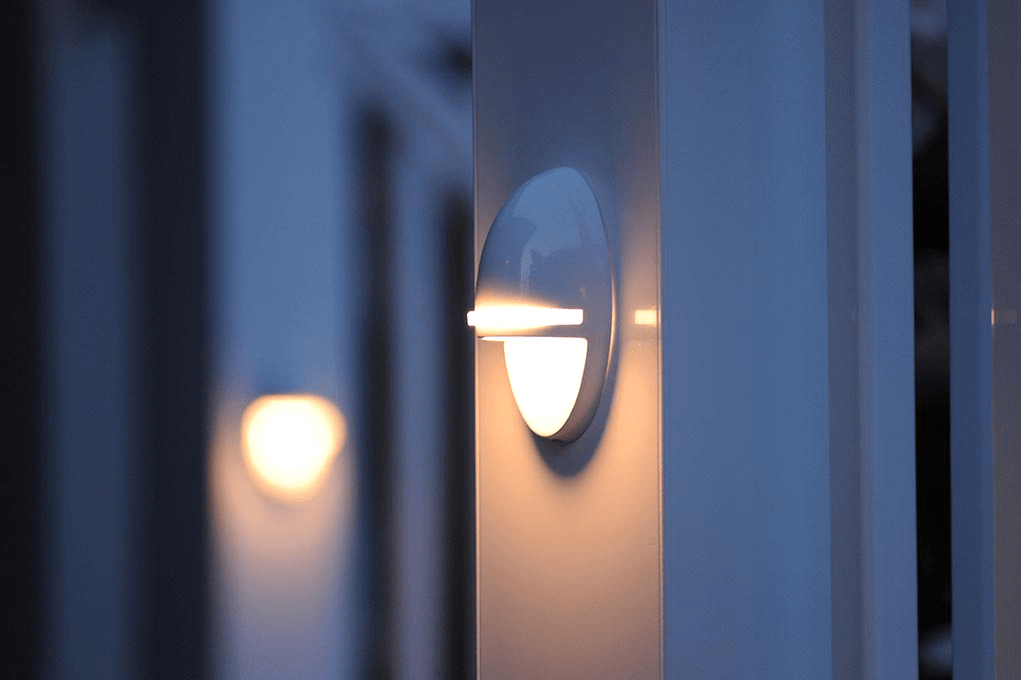 Two round, lit sconces on a white fence, one close and in focus, the other blurry in the background.