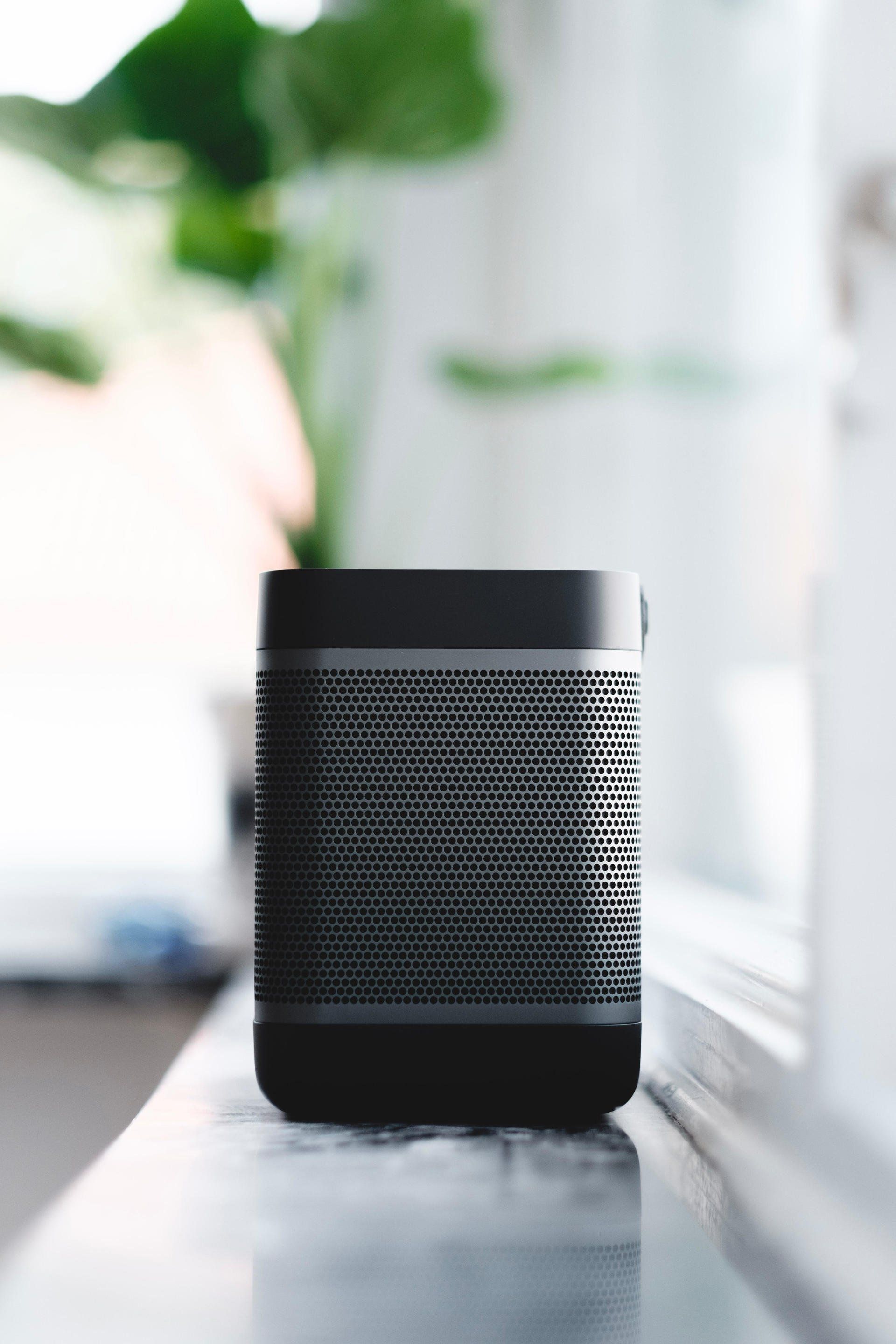 Black portable speaker on a white windowsill, with a blurry green plant visible in the background.