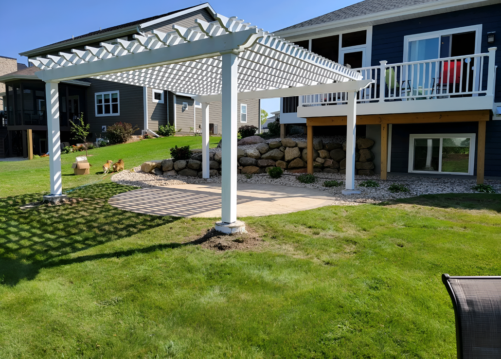 A white vinyl pergola stands on a gravel and stone patio in a backyard, positioned next to a dark blue house with a deck.