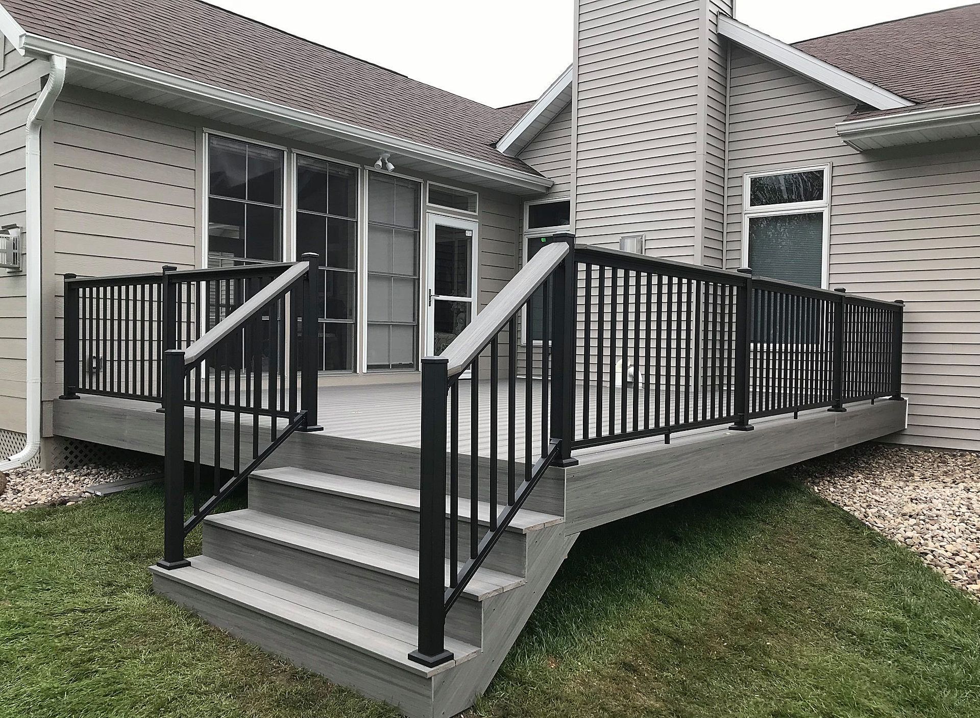 A light gray composite deck with black metal railings and a staircase, attached to the back of a beige residential home.