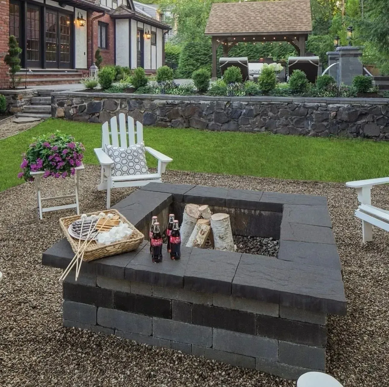 Fire pit with firewood, chairs, snacks on gravel patio with a house and garden in the background.