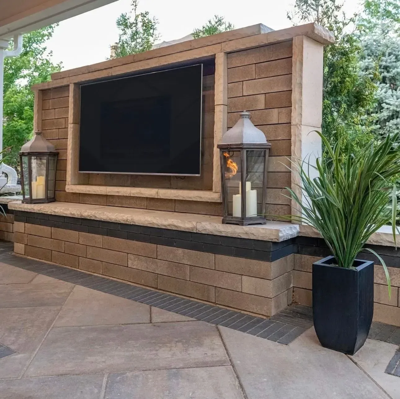 Outdoor TV mounted in a brick enclosure with lanterns and a potted plant.