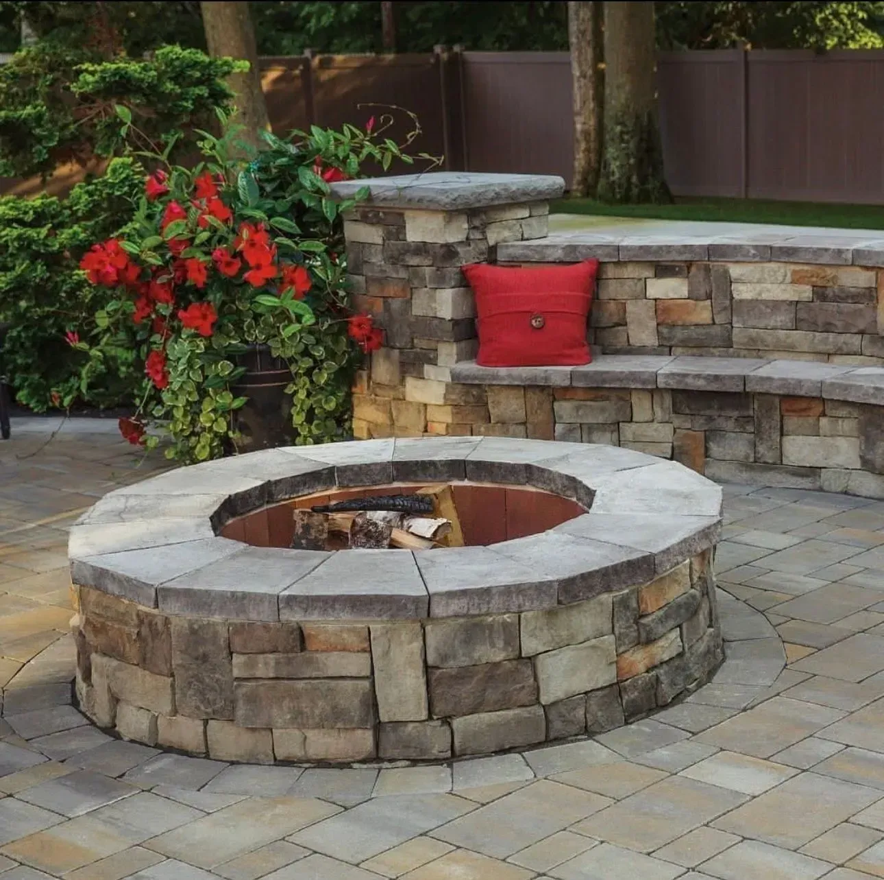 Stone fire pit with built-in seating; red cushion, red flowers, and brick patio.
