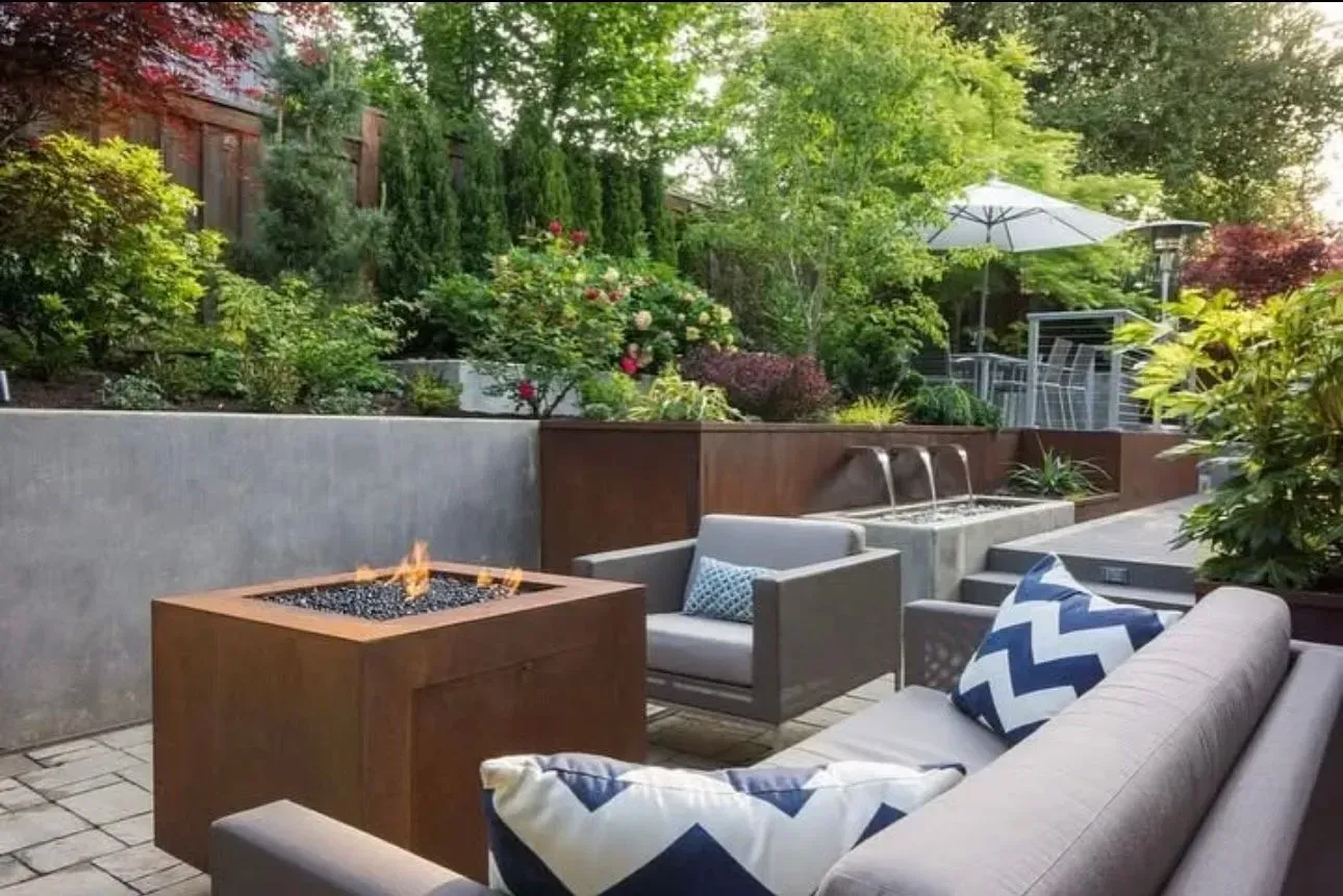 Patio with a modern fire pit, seating, and a water feature surrounded by lush greenery.