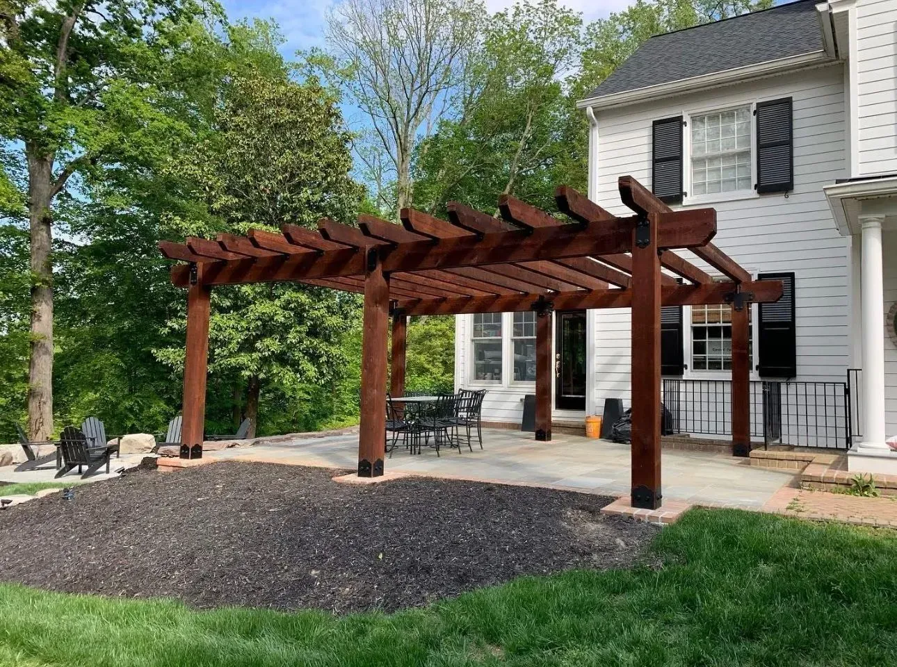 Wooden pergola over a patio next to a two-story house. Dark brown, with a dining set inside and lush greenery surrounding.