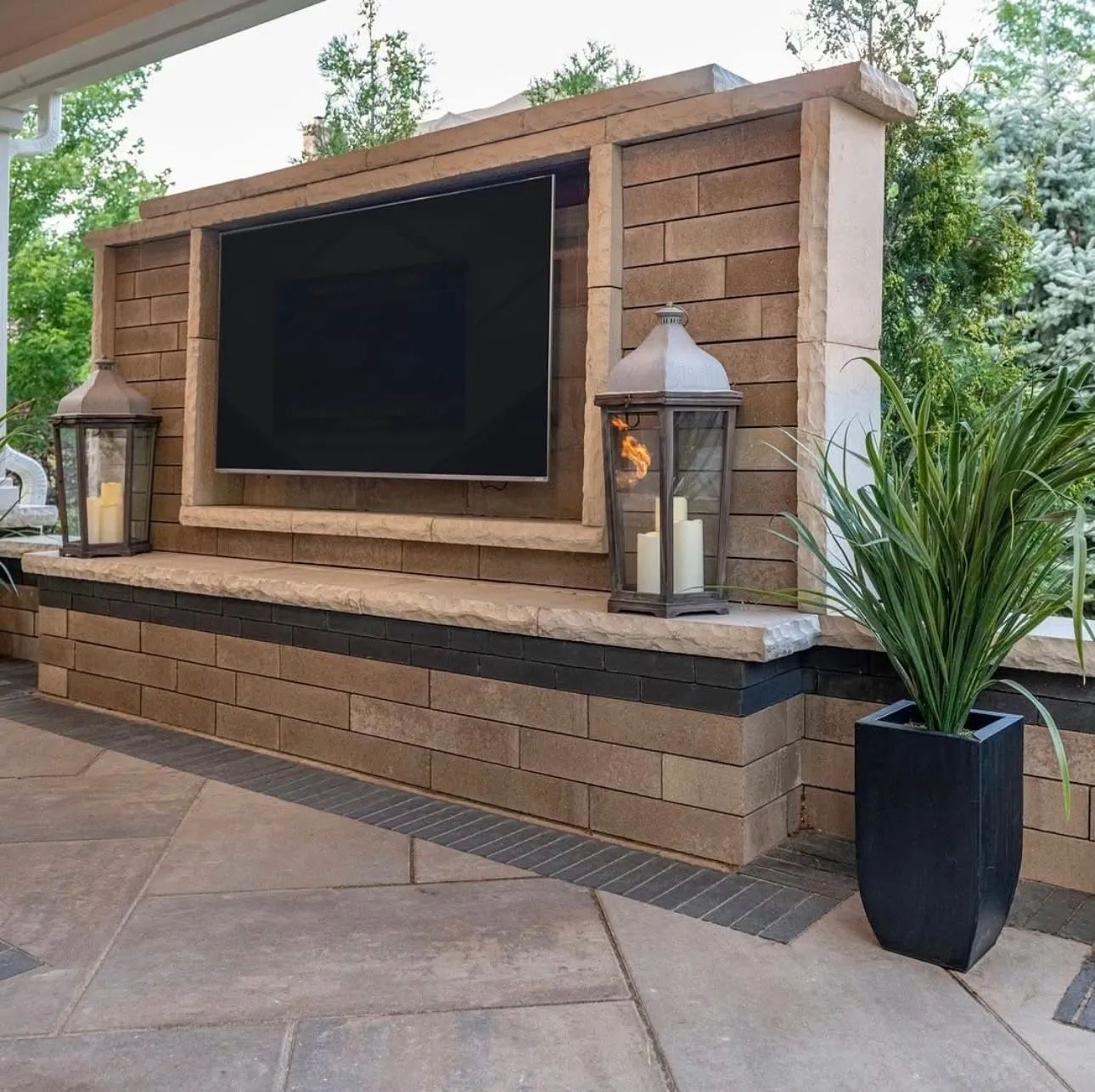 Outdoor TV built into a stone wall with lanterns and a potted plant on a patio.