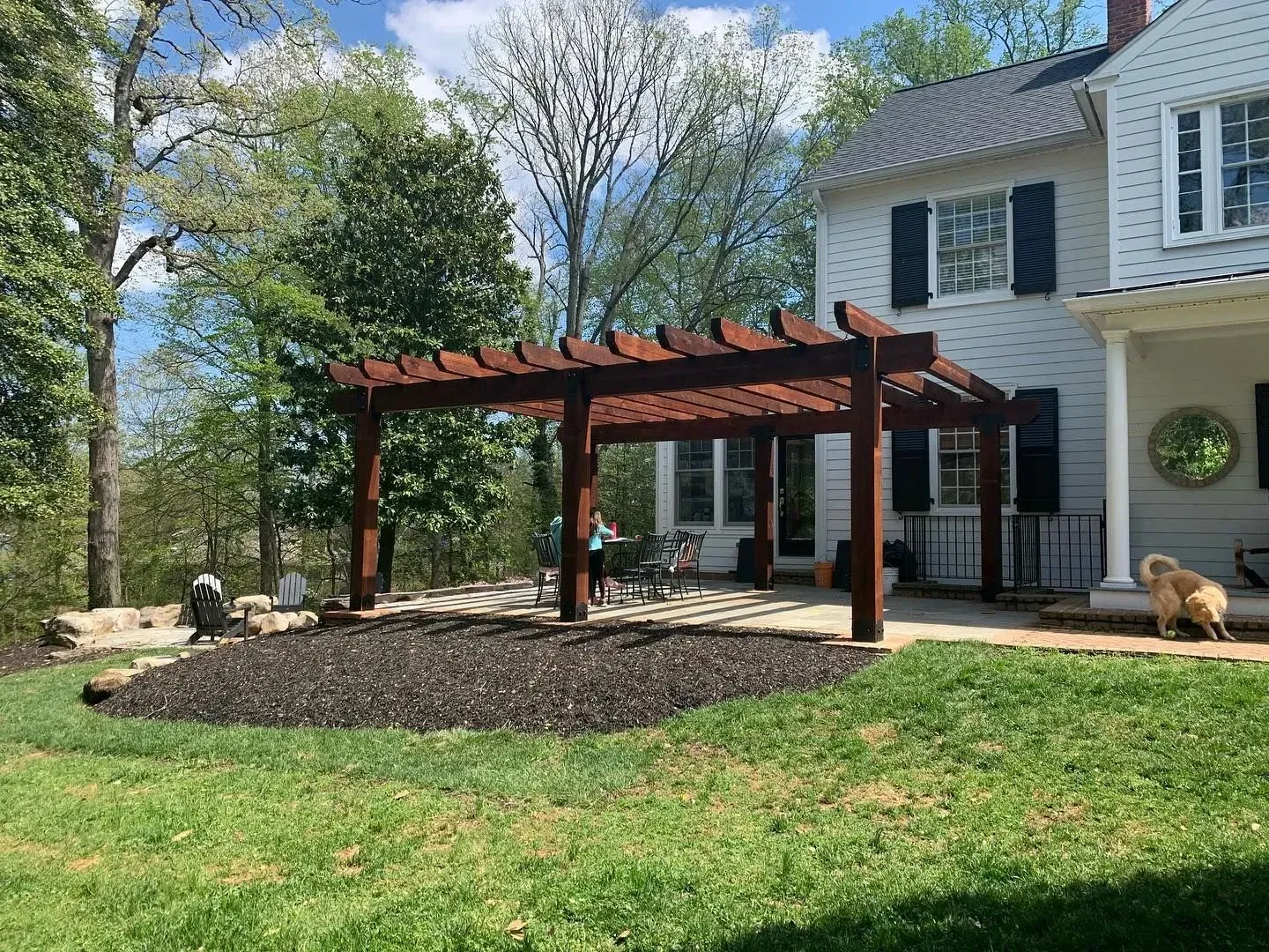 Pergola with brown beams and posts near a white house with black shutters.