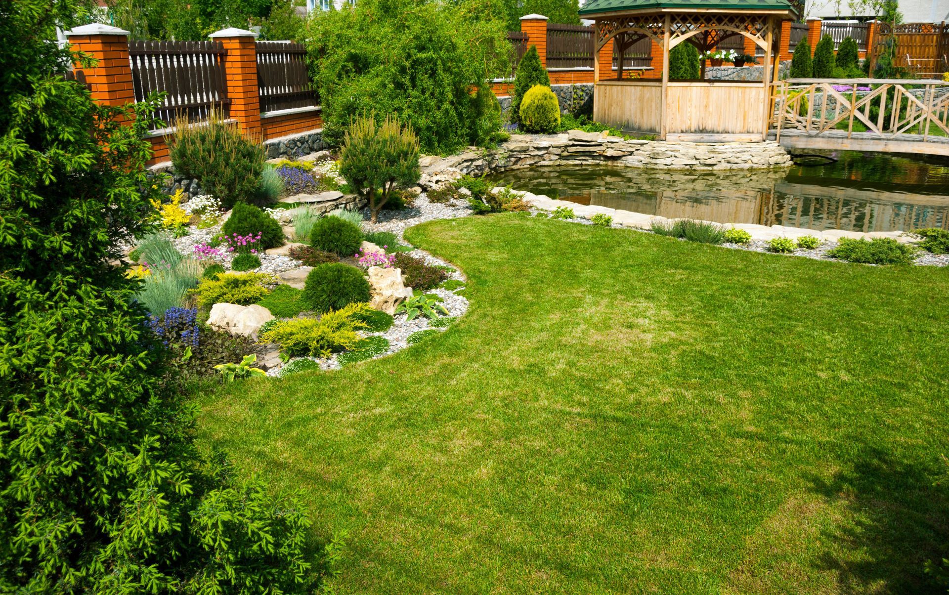 Lush green lawn with a landscaped garden, pond, and wooden gazebo in a sunny backyard.