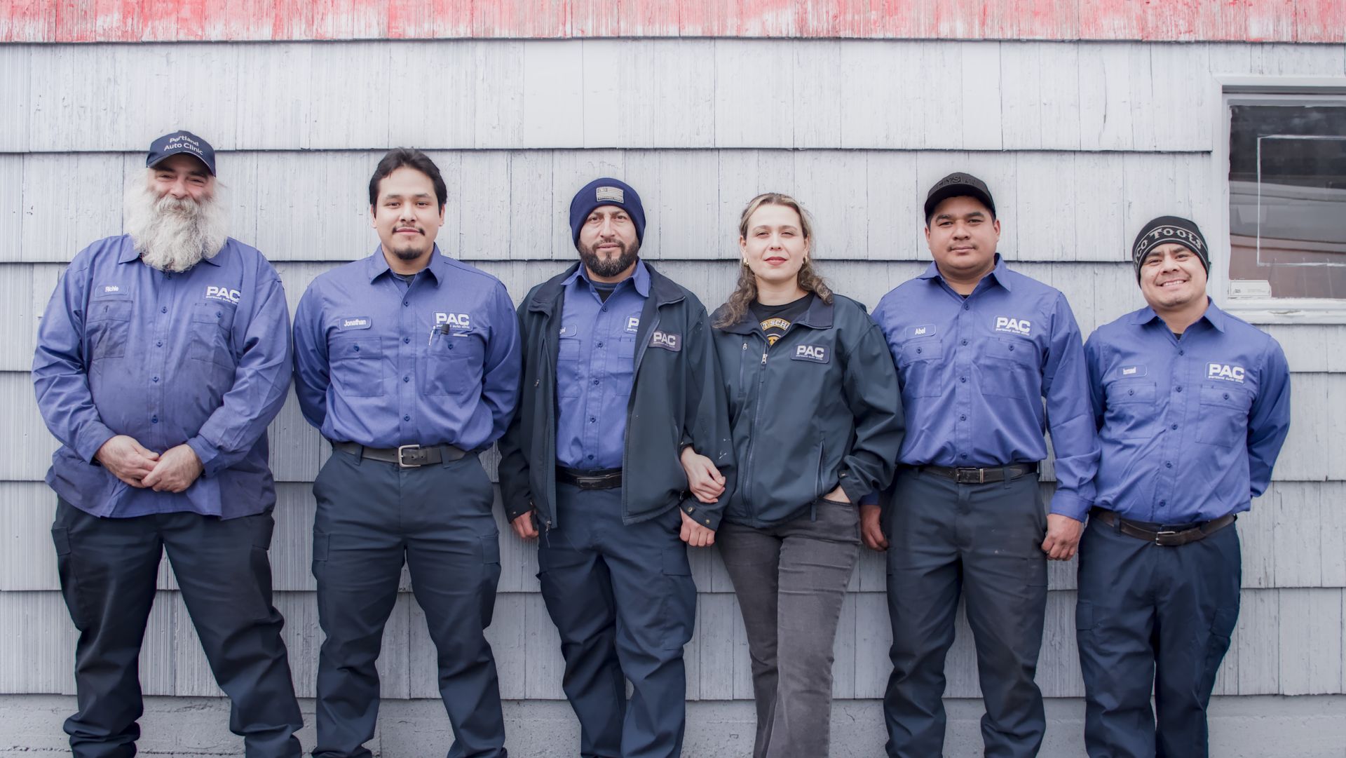 Six people in matching blue shirts and pants stand in front of a gray building.