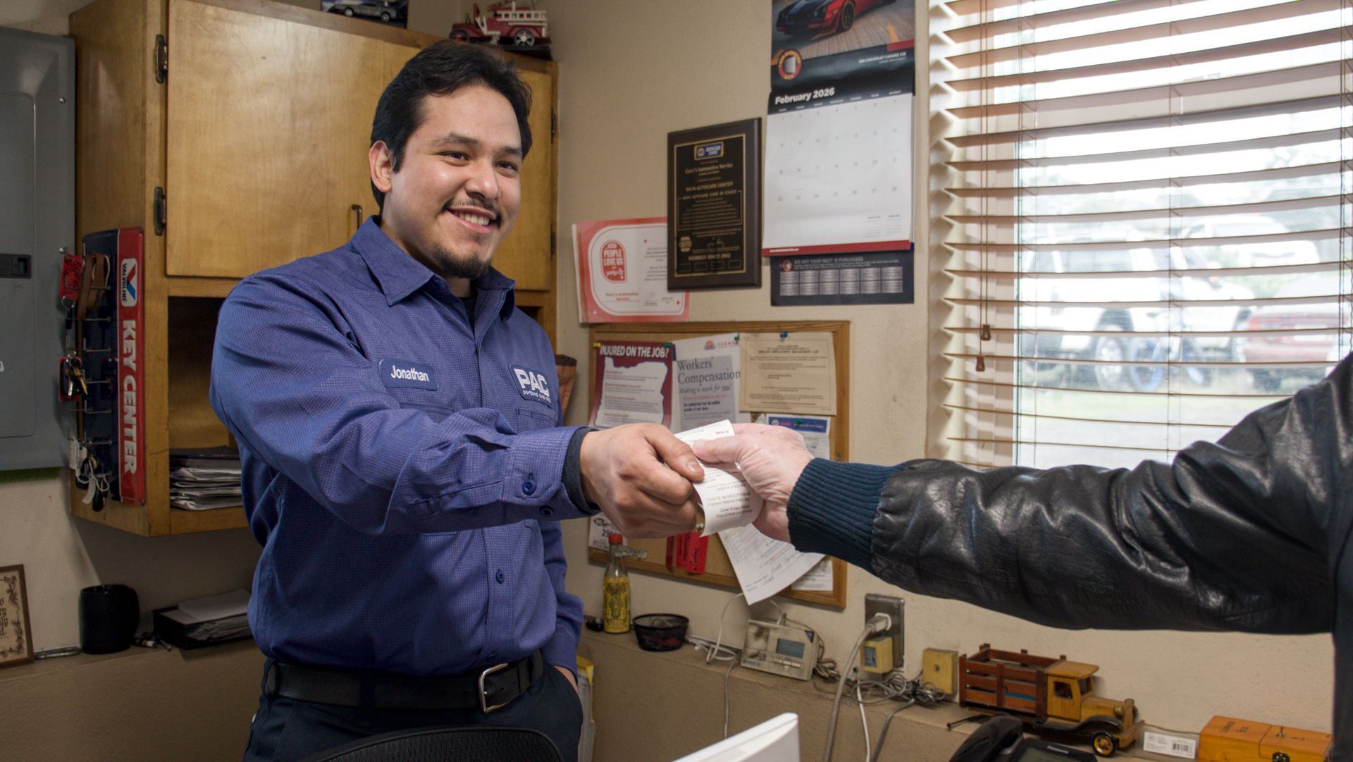Mechanic in blue shirt handing something to a person, likely a customer, inside a small office.