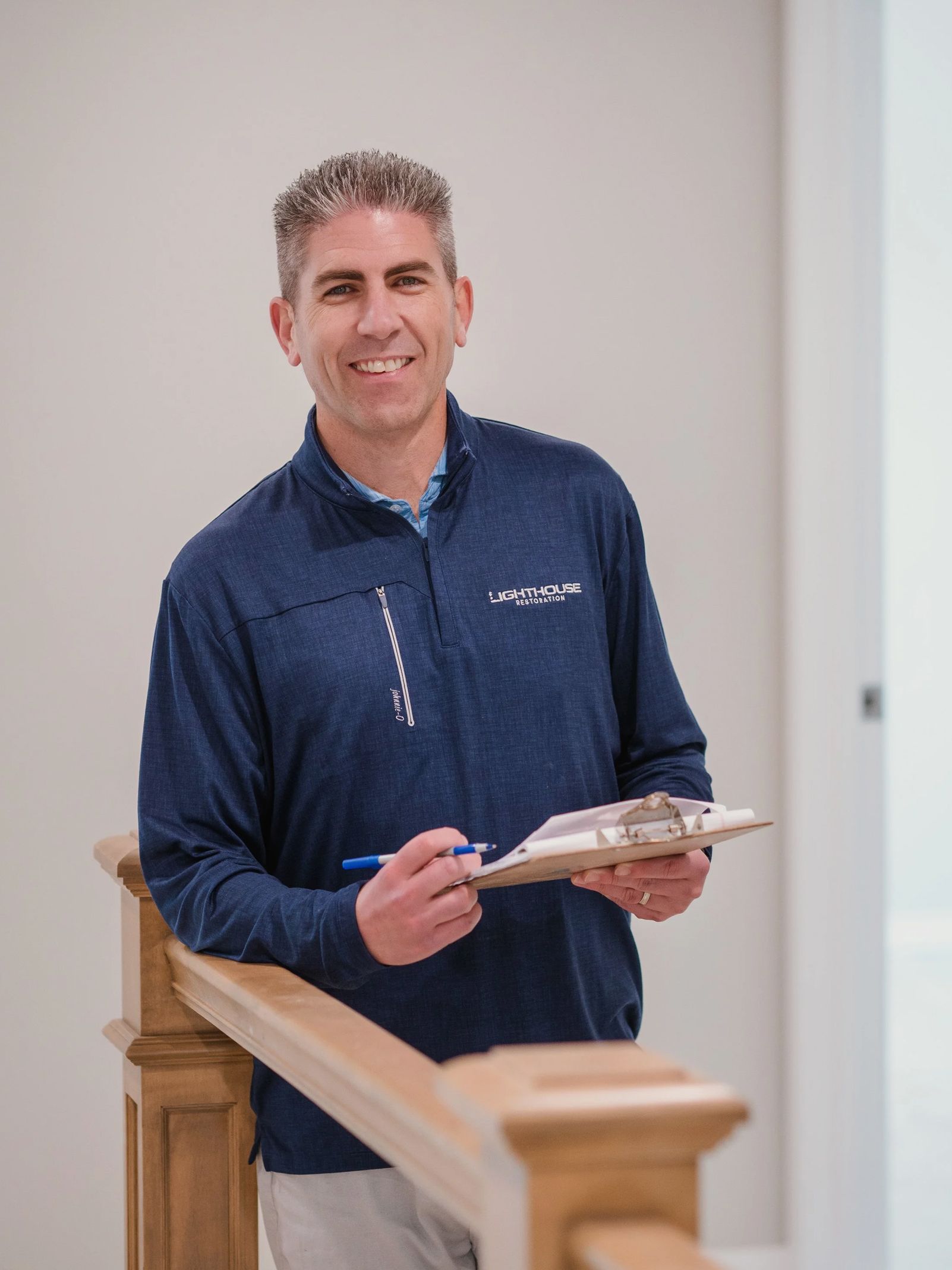 Man holding a clipboard while standing by a stair railing indoors, wearing a navy shirt and smiling