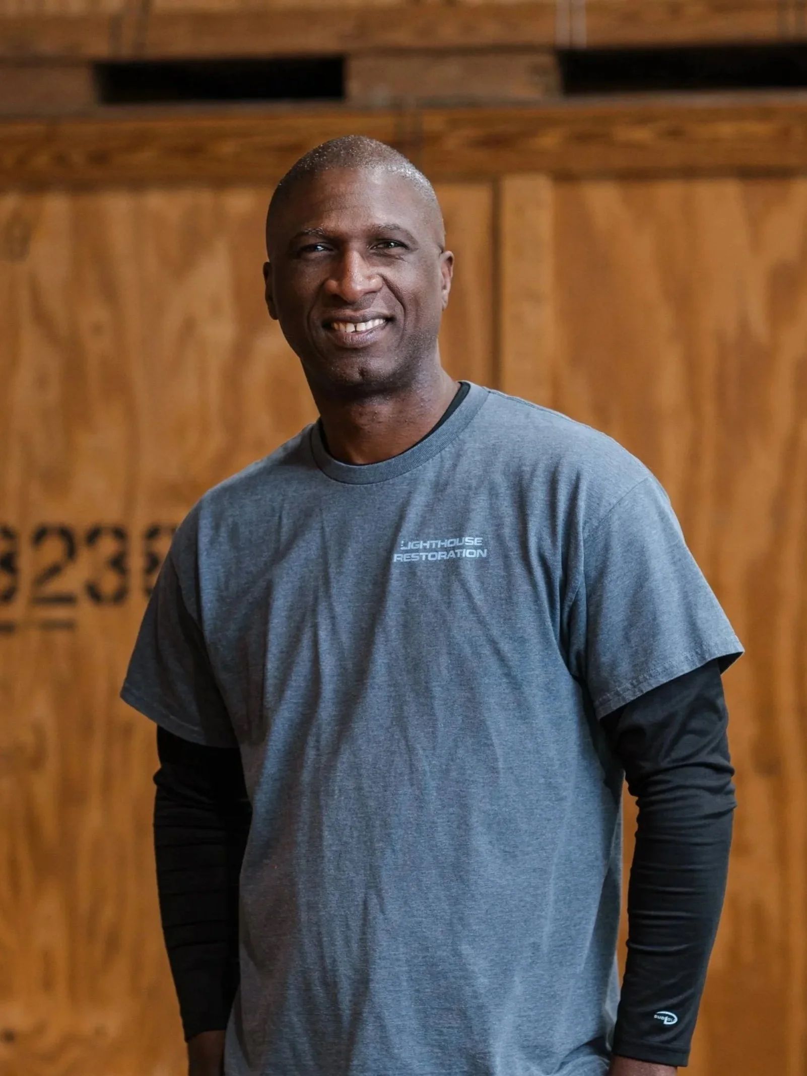 Man standing indoors in a gray T-shirt and black long sleeves against a wooden wall.