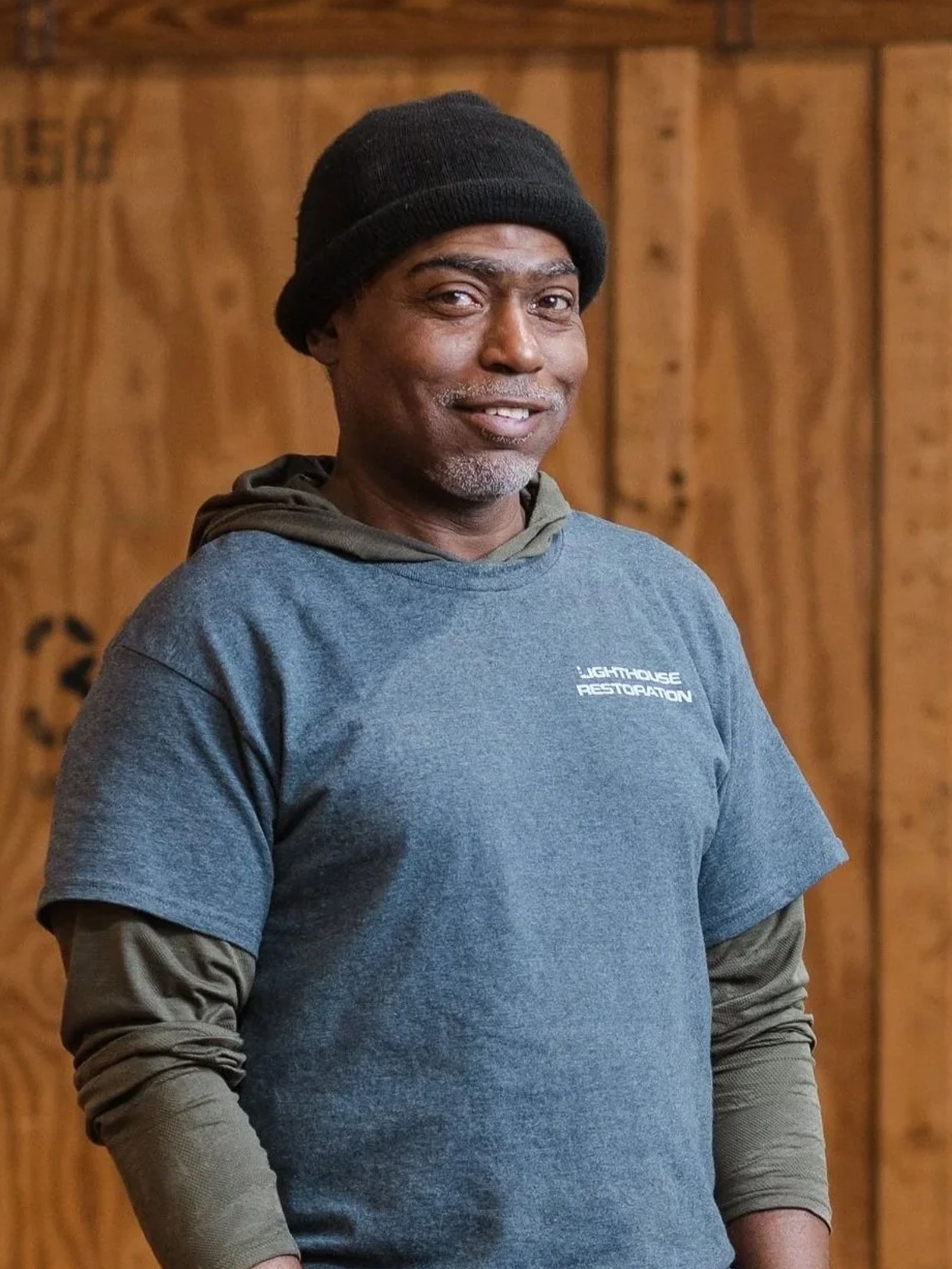Person in a black beanie and blue T-shirt stands indoors against a wooden wall, looking at the camera.