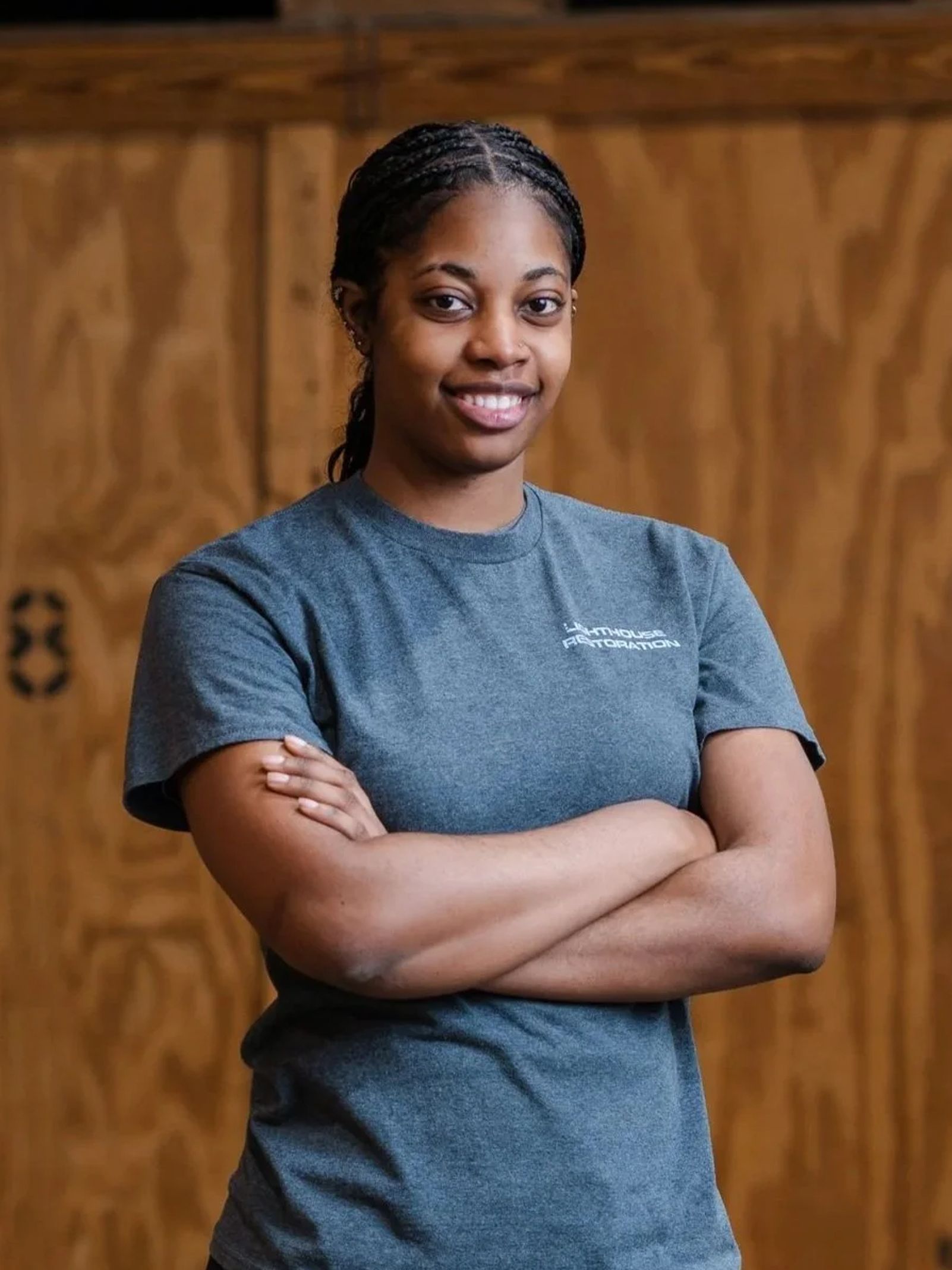 Person standing with arms crossed in a gray T-shirt against a wooden wall.