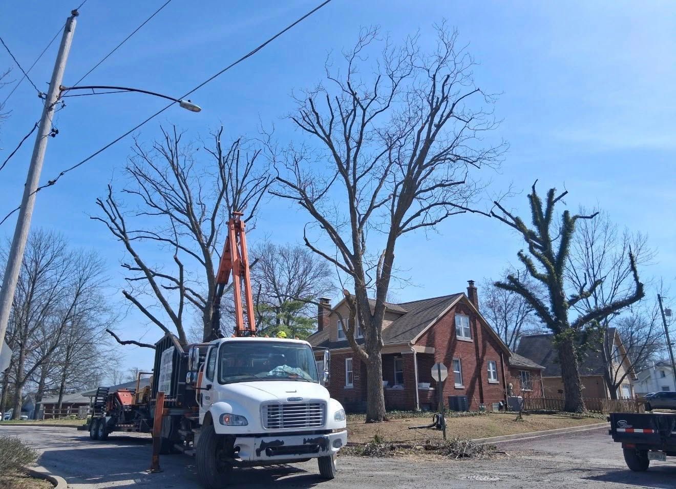 Utility truck with boom lift beside a brick house and leafless trees on a sunny street.