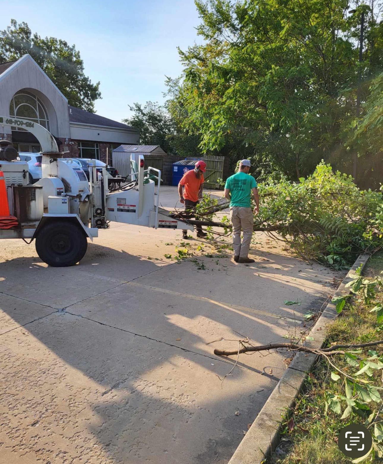 Tree service workers using a chipper machine to process branches, in front of a building.