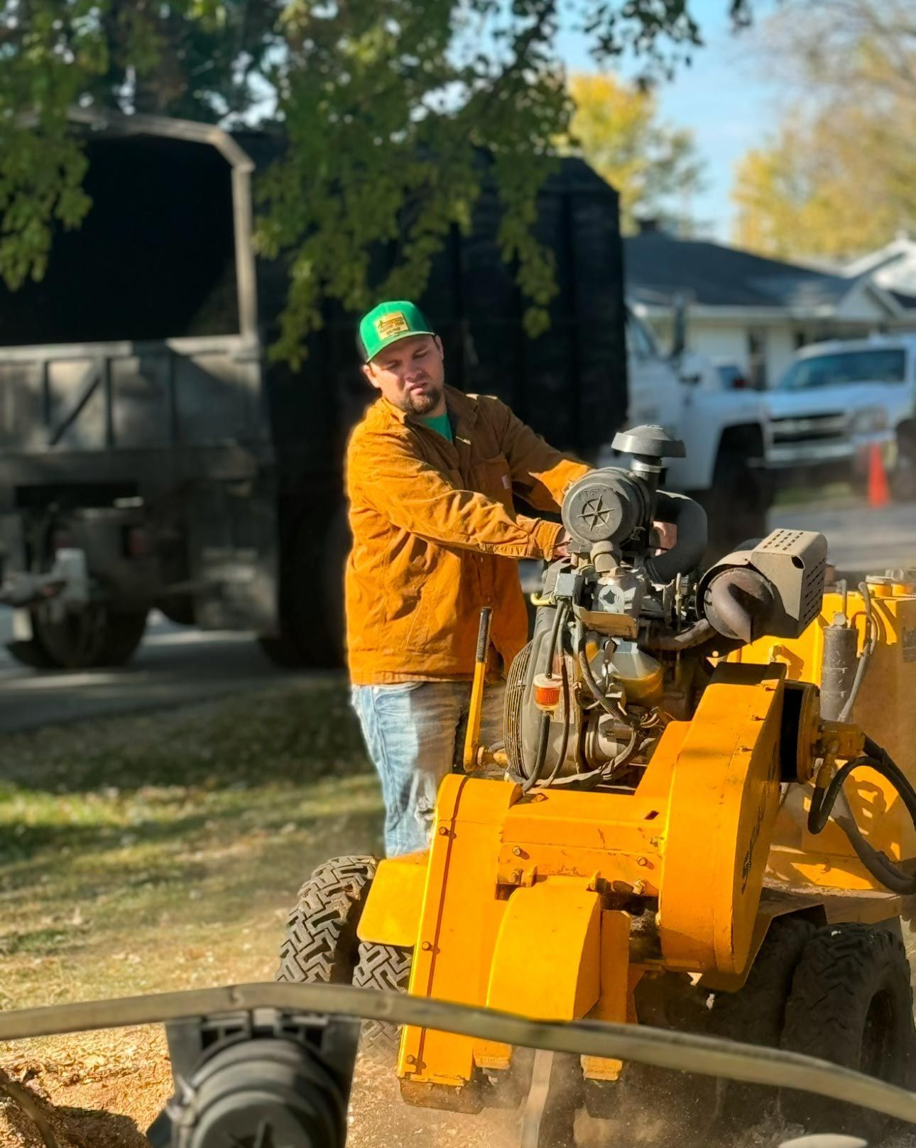 Man operating a yellow stump grinder on a street. He wears a gold jacket and green hat. A truck is in the background.