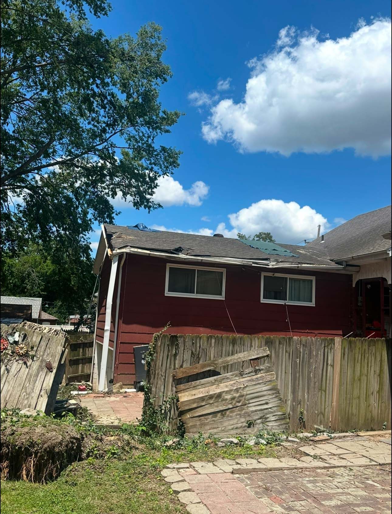 Damaged red house with missing roof shingles and a dilapidated wooden fence under a blue sky with clouds.