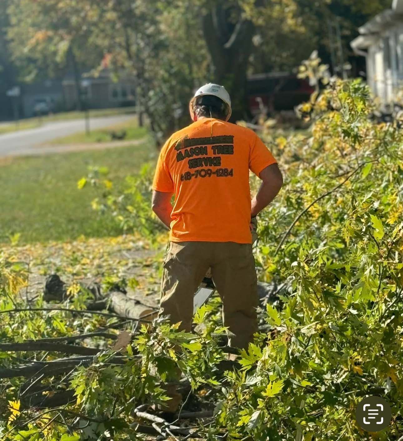 Man in orange shirt with tree service logo standing amidst tree debris. Outdoors on a sunny day.