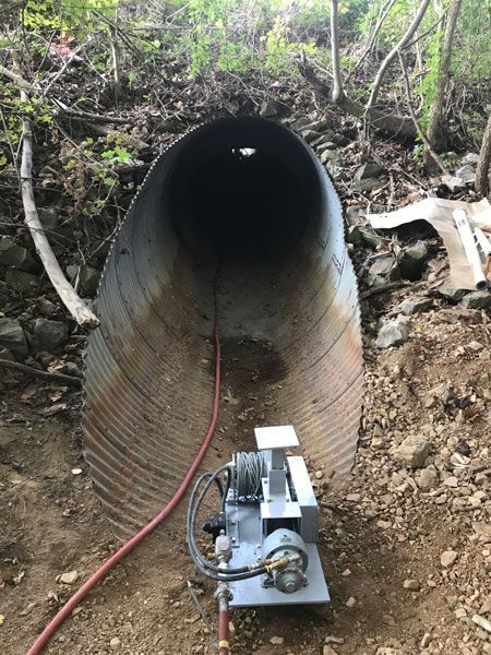 Large concrete pipe tunnel entrance with a machine and red hose at the base in a dirt embankment
