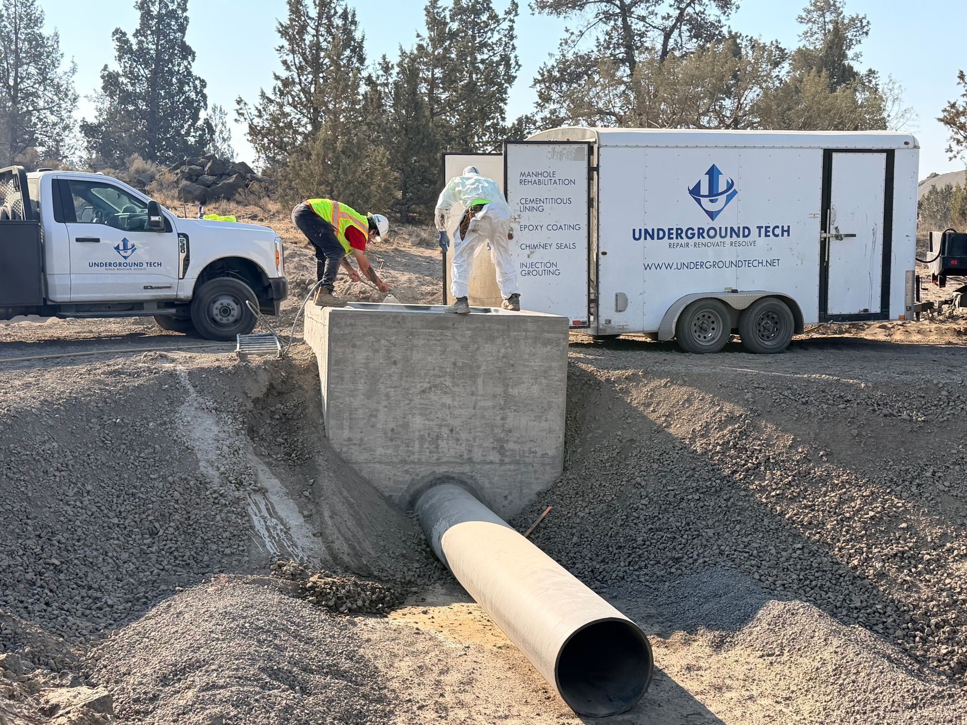 Workers install a large pipe at a roadside drainage site beside trucks and equipment.