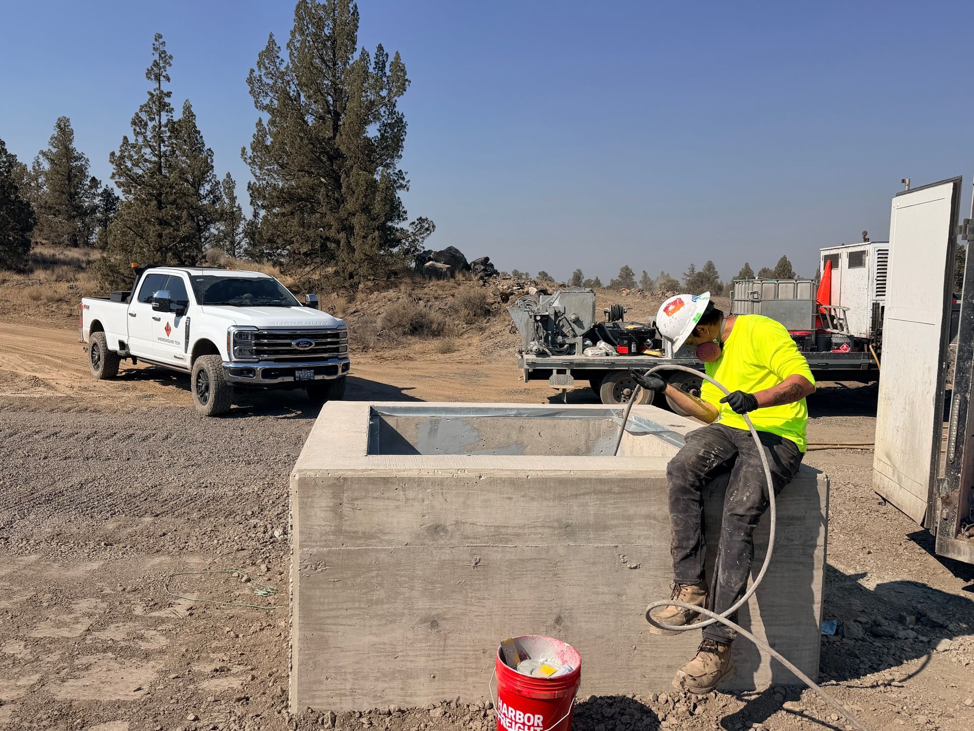 Worker in neon jacket using a hose beside a concrete tank at a rural construction site with trucks nearby